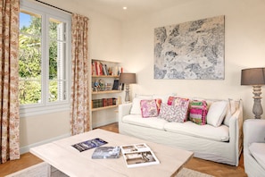 A living room with a white sofa, a coffee table with magazines, a bookshelf, and a large abstract painting on the wall. - Mas des Cigales – Pool & Saint-Paul views (Vence)
