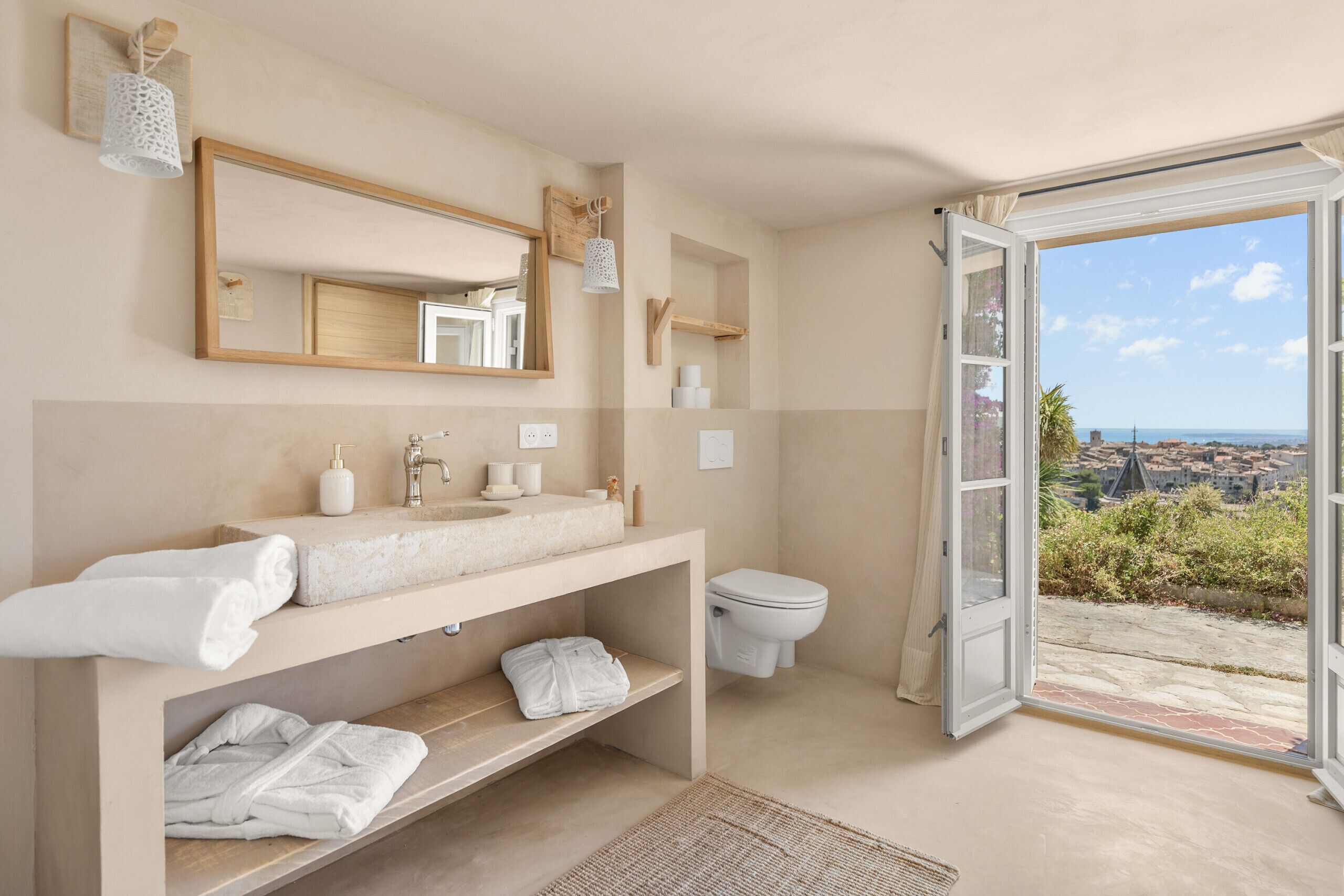 A bathroom with a large mirror, a sink, a toilet, and a view of the city through an open door.
