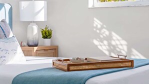 Ein ordentlich bezogenes Bett mit blauer Decke, einer Nachttischlampe, einem kleinen Topf mit Pflanze und einem Tablett mit Buch und Schale.