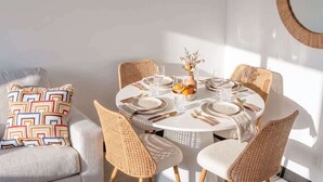 Ein runder Esstisch mit sechs Korbstühlen, ein weißes Sofa mit einem gemusterten Kissen und ein Spiegel an der Wand.