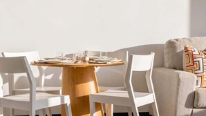 Comedor con una mesa de madera redonda, sillas blancas y un sofá beige con un cojín estampado.