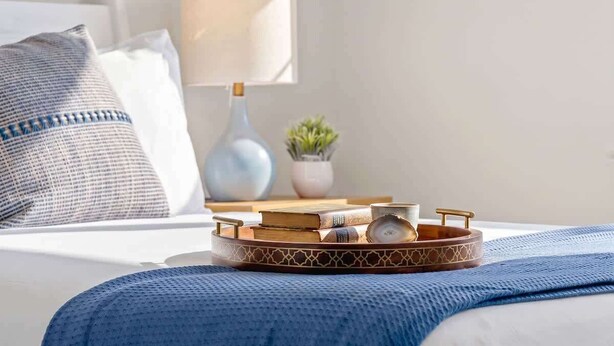 Una cama bien tendida con una manta azul, una mesita de noche con una lámpara, una planta pequeña y una bandeja con libros y una taza.