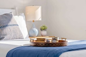 Een net opgemaakt bed met een blauwe deken, een nachtkastje met een lamp, een kleine plant en een dienblad met boeken en een beker.