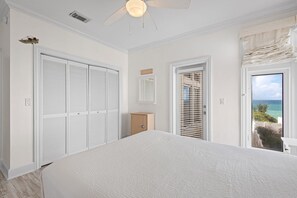 A bedroom with a large bed, a ceiling fan, a view of the ocean, and a closet with white shutters.