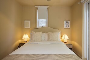 A bedroom with a bed, two bedside lamps, a window with a blind, and two framed pictures on the wall. - Blythe & Bloom Garden Flat (London)