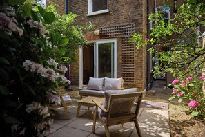 A patio with a sofa and chairs, a brick wall, and a window. - Blythe & Bloom Garden Flat (London)