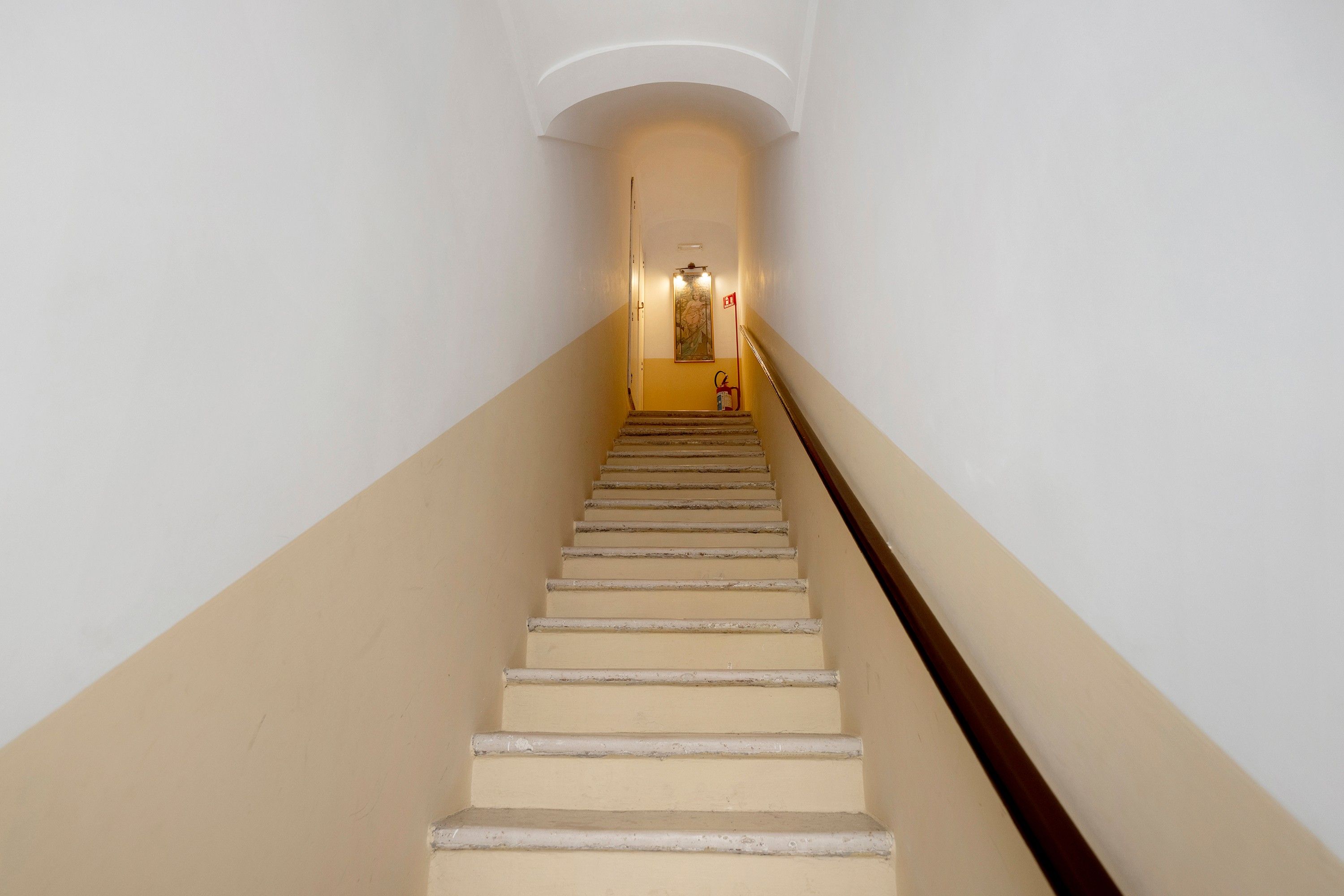 Eine Treppe mit Handlauf führt zu einer Zwischenebene, auf der sich eine Leuchte und ein Feuerlöscher befinden.