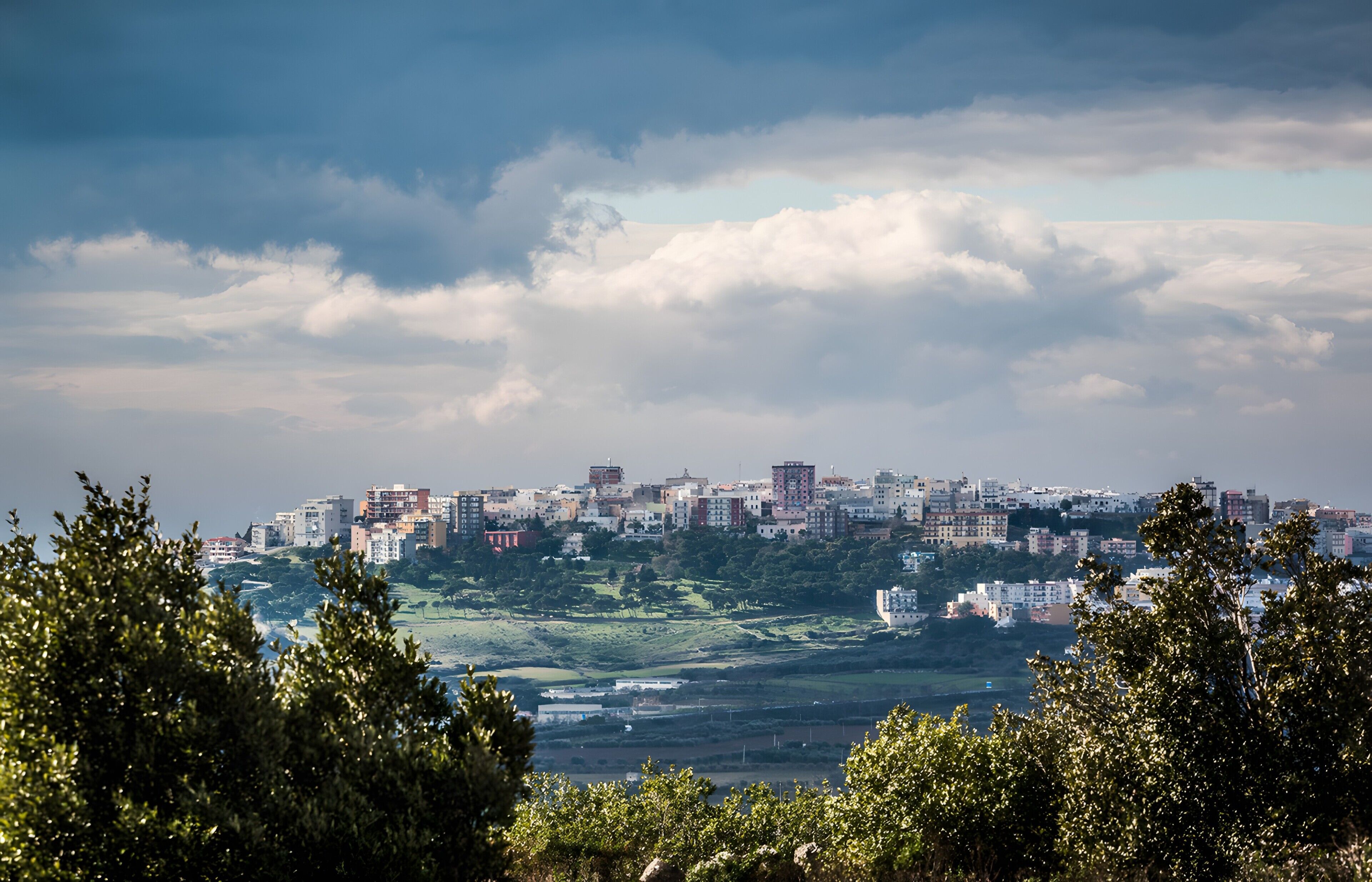 Une vue urbaine avec des bâtiments situés sur une colline, entourés d’arbres et de nuages.