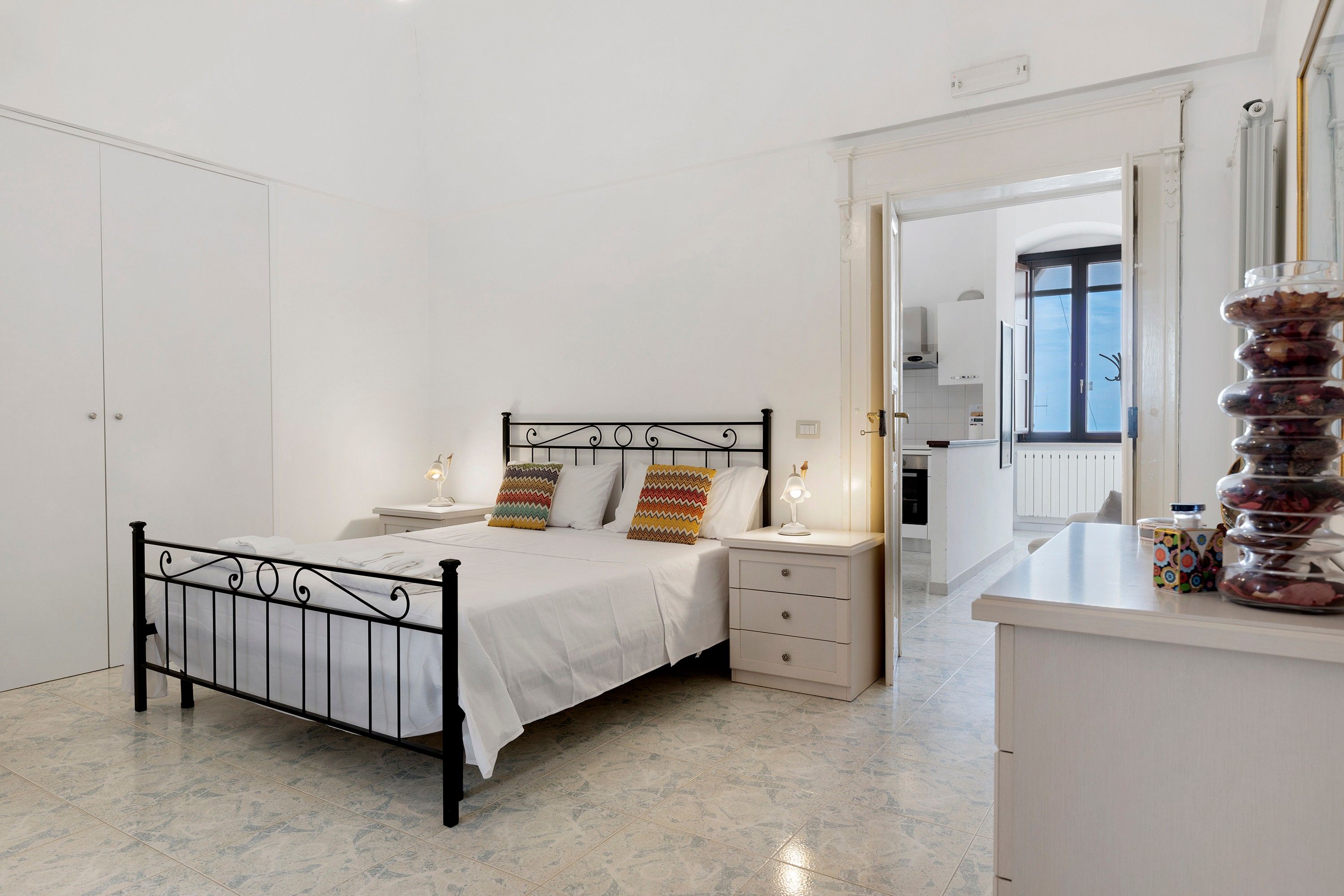 Ein Schlafzimmer mit einem Bett, Nachttischen, einem Spiegel und Blick zur Küche.