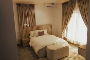 A modern bedroom with a large bed, a wooden headboard, a bedside table, a nightstand, and a window with curtains.