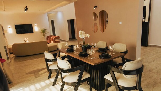 A dining area with a wooden table set for six, surrounded by six chairs with black frames and white cushions. - The Embassy Boutique Apartment (Jos)