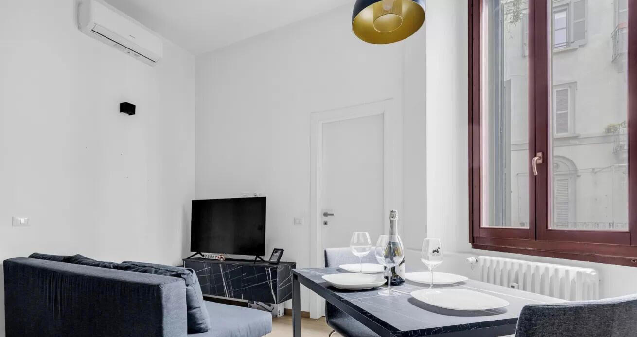 A modern living room with a sofa, a TV, a dining table set for two with wine glasses, and a window with a view of buildings.