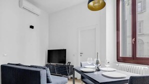 Un salon moderne comprenant un canapé, un téléviseur, une table à manger dressée pour deux personnes avec des verres à vin, et une fenêtre donnant sur des immeubles.