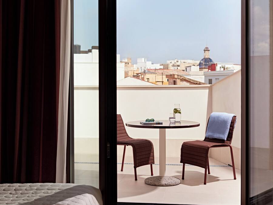 A room with a bed, a balcony with two chairs, a table, and a view of buildings.