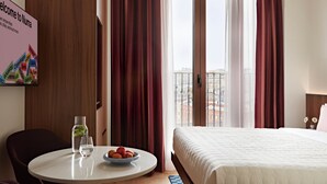 A hotel room with a bed, a round table with a bowl of fruit, a chair, and a view of the city through large windows.