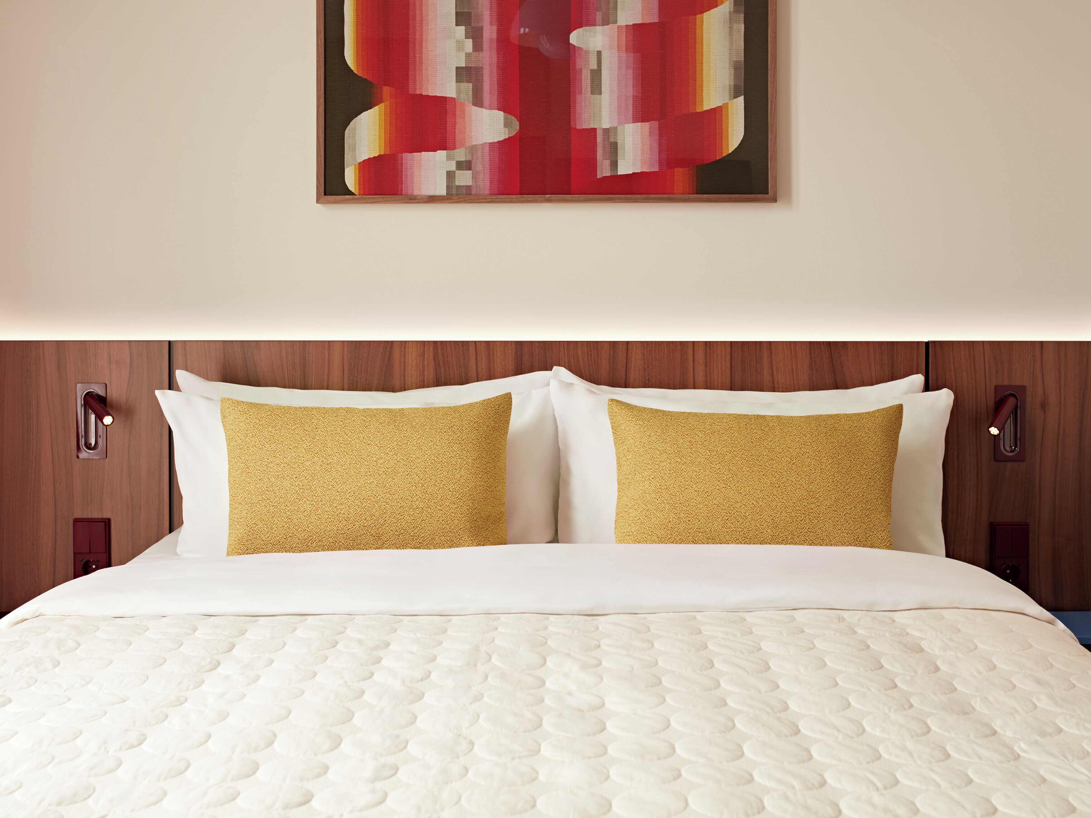 A neatly made bed with a white quilt and two pillows, a wooden headboard, and a wall-mounted abstract painting.