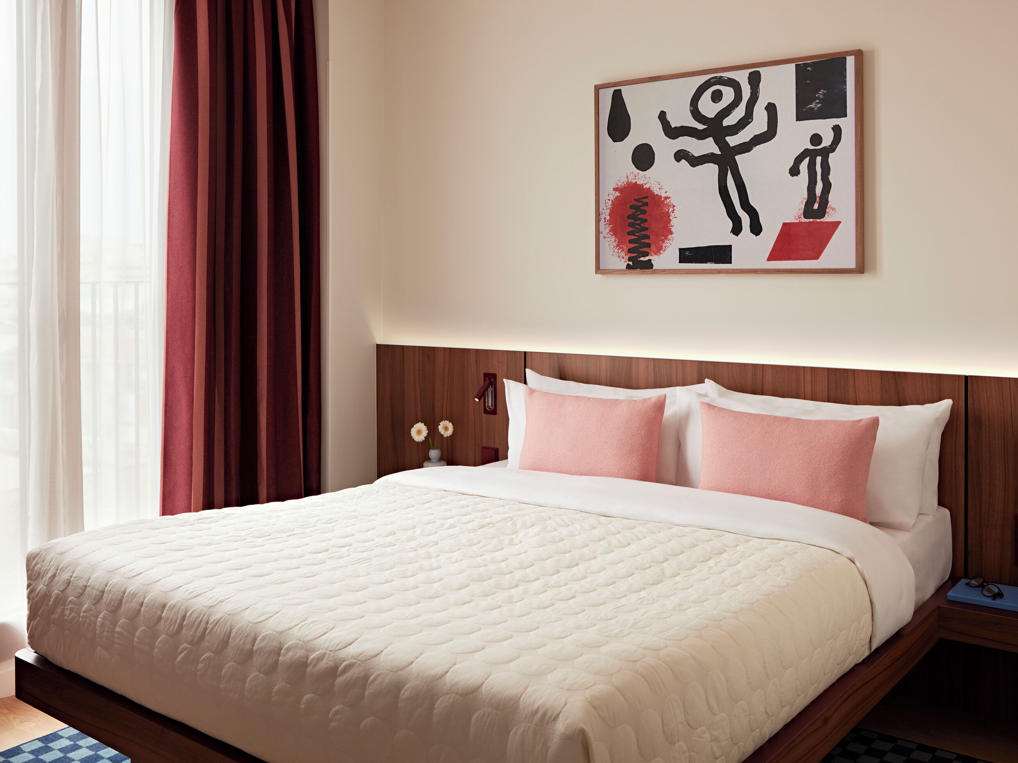 A hotel room with a bed, a nightstand, a framed artwork, and a window with curtains.