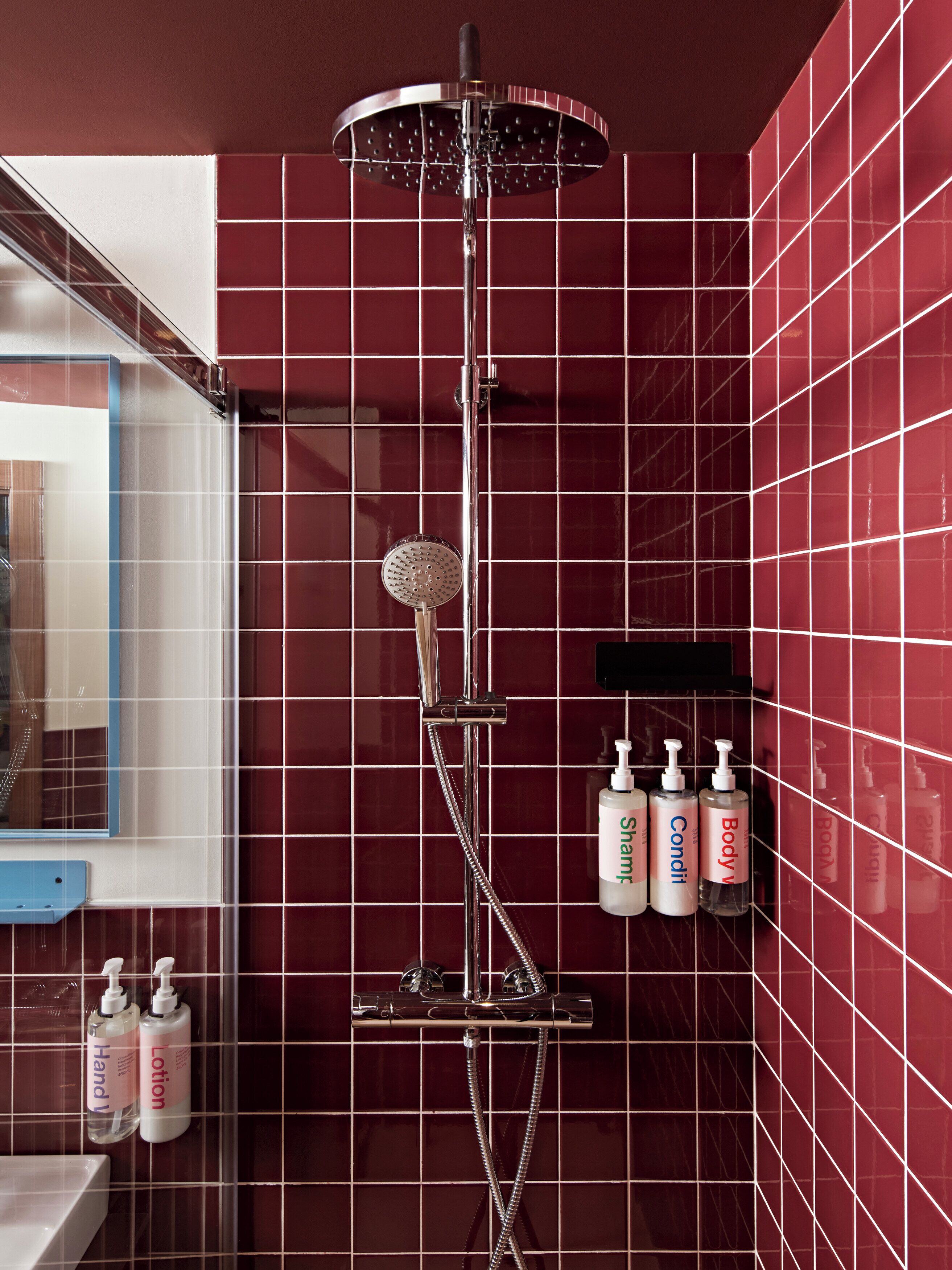 Un baño con ducha vertical, paredes de azulejos rojos y artículos de aseo en un estante.