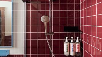 Un baño con ducha vertical, paredes de azulejos rojos y artículos de aseo en un estante.
