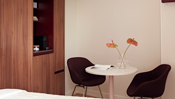 A hotel room with a bed, a wooden wardrobe, a small table with two chairs, and a vase with flowers.