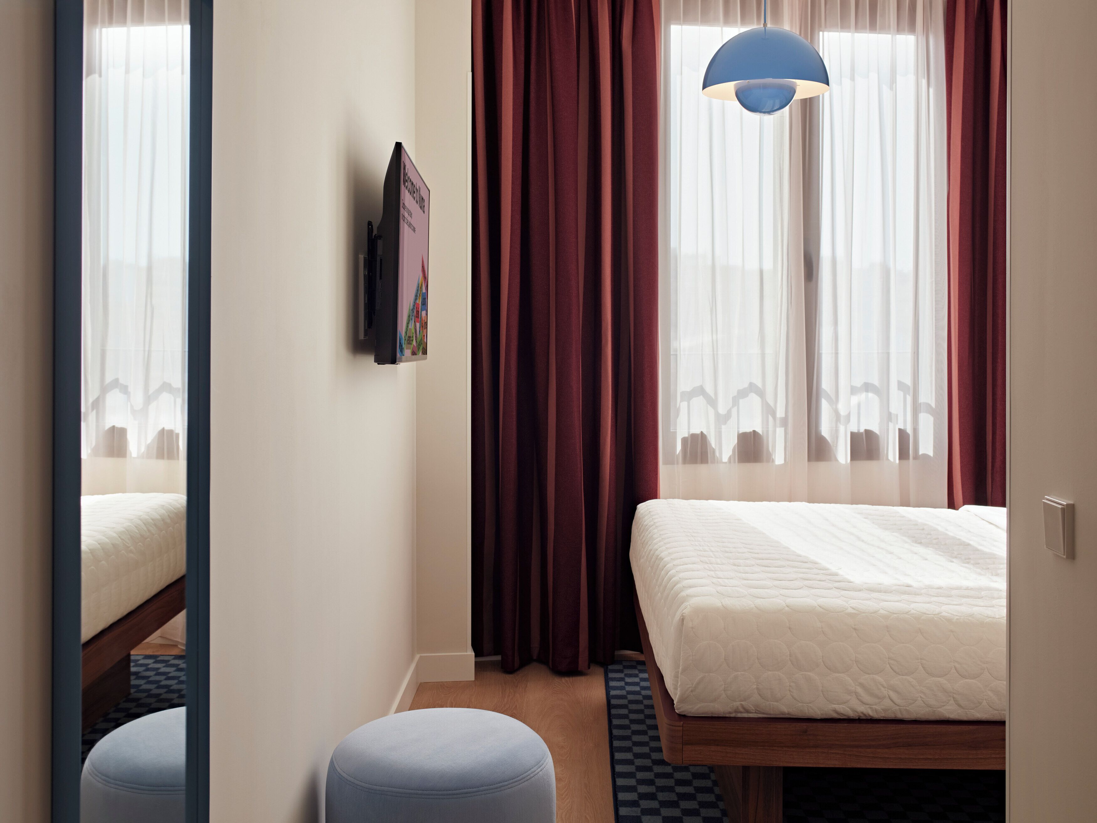 A hotel room with a bed, a blue ottoman, a wall-mounted TV, and a window with curtains.
