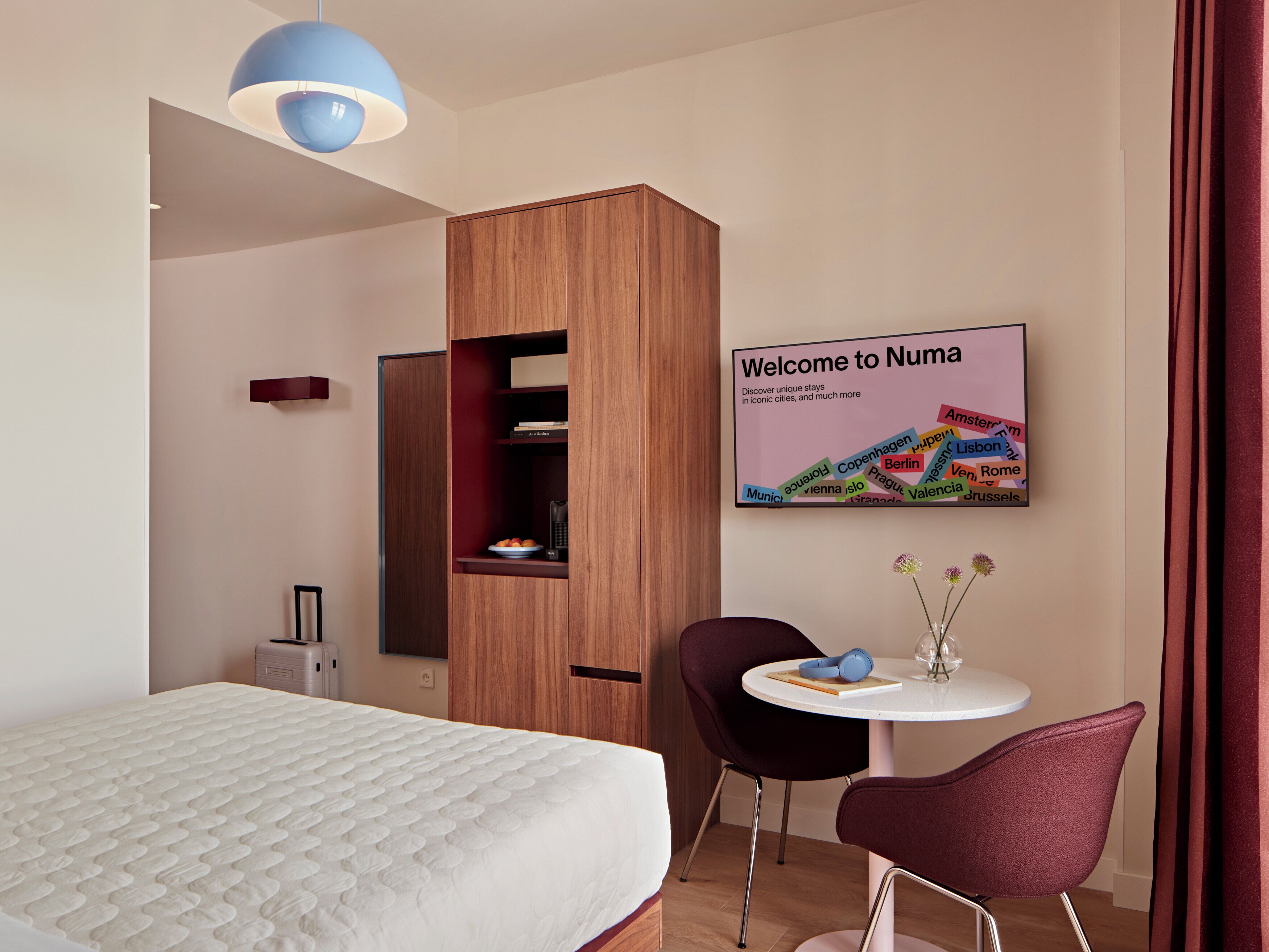Ein Hotelzimmer mit Bett, Schrank, kleinem Tisch mit Stuhl, einem Wandzeichen und einer blauen Pendelleuchte.
