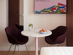 A hotel room with a round table, two chairs, a bed, and a wall with a colorful city names poster.