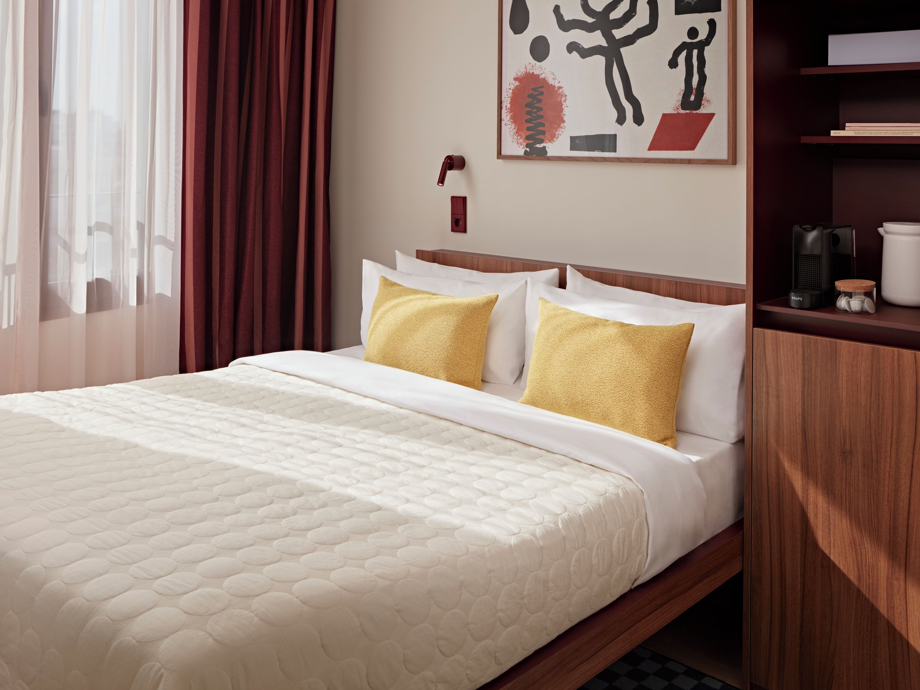 A hotel room with a bed, a nightstand, a coffee maker, and a painting on the wall.