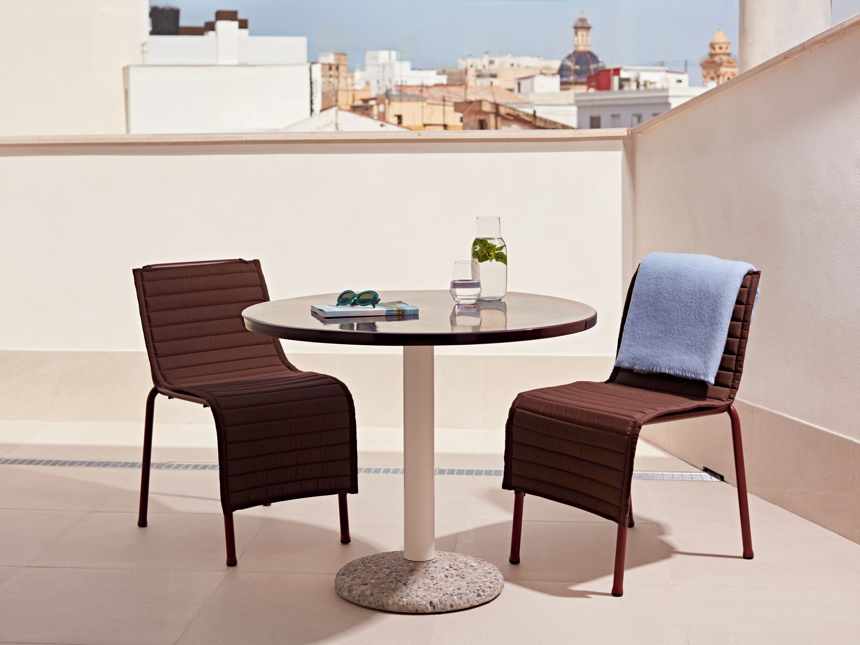 Una mesa redonda con dos sillas en un balcón, un florero con una planta y un par de gafas de sol sobre la mesa.