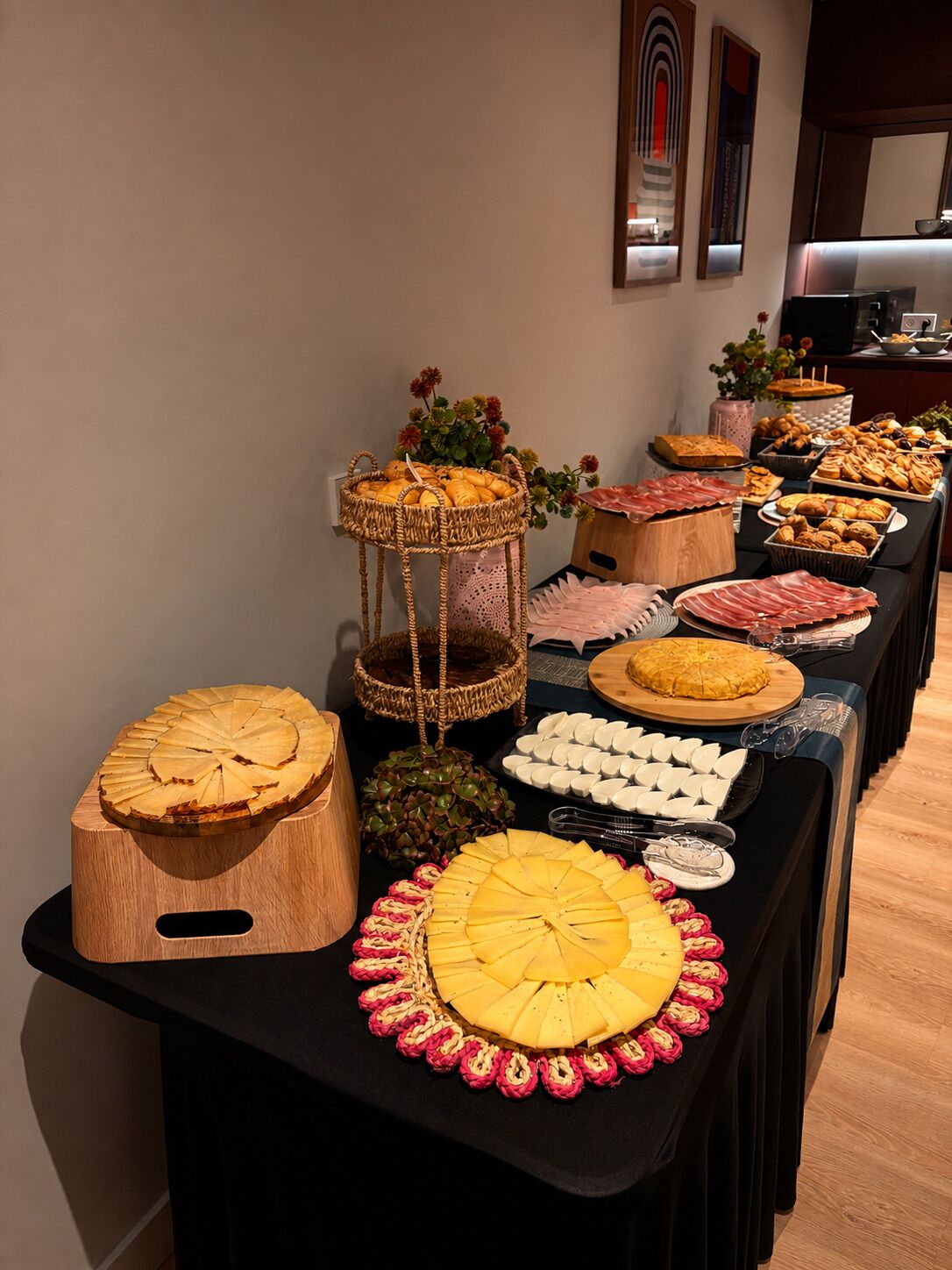 Una mesa de buffet con diversos alimentos, incluyendo una tabla de madera grande con un diseño circular, una cesta con flores y una bandeja con objetos blancos y redondos.