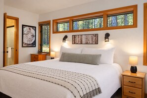 A bedroom with a large bed, two bedside tables, a nightstand, and a window with a view of trees. - Luxury 5BR Forest Retreat at Black Butte Ranch (Sisters)
