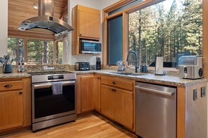 A modern kitchen with wooden cabinets, stainless steel appliances, a built-in microwave, and a large window offering a view of a forest. - Luxury 5BR Forest Retreat at Black Butte Ranch (Sisters)