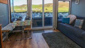 Une pièce chaleureuse avec une table à manger dressée pour deux personnes, un canapé agrémenté de coussins et de grandes fenêtres offrant une vue sur un paysage verdoyant.