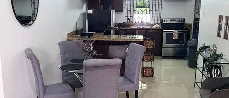 A modern kitchen with dark cabinets, a central island, and a dining area with grey chairs.