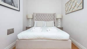 Un lit bien fait, avec du linge de lit blanc et une tête de lit capitonnée, deux tables de chevet avec des lampes et un tableau encadré au mur.