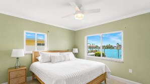 Une chambre à coucher avec un lit, deux tables de chevet avec des lampes, un ventilateur de plafond et une vue sur une étendue d’eau grâce à de grandes fenêtres.