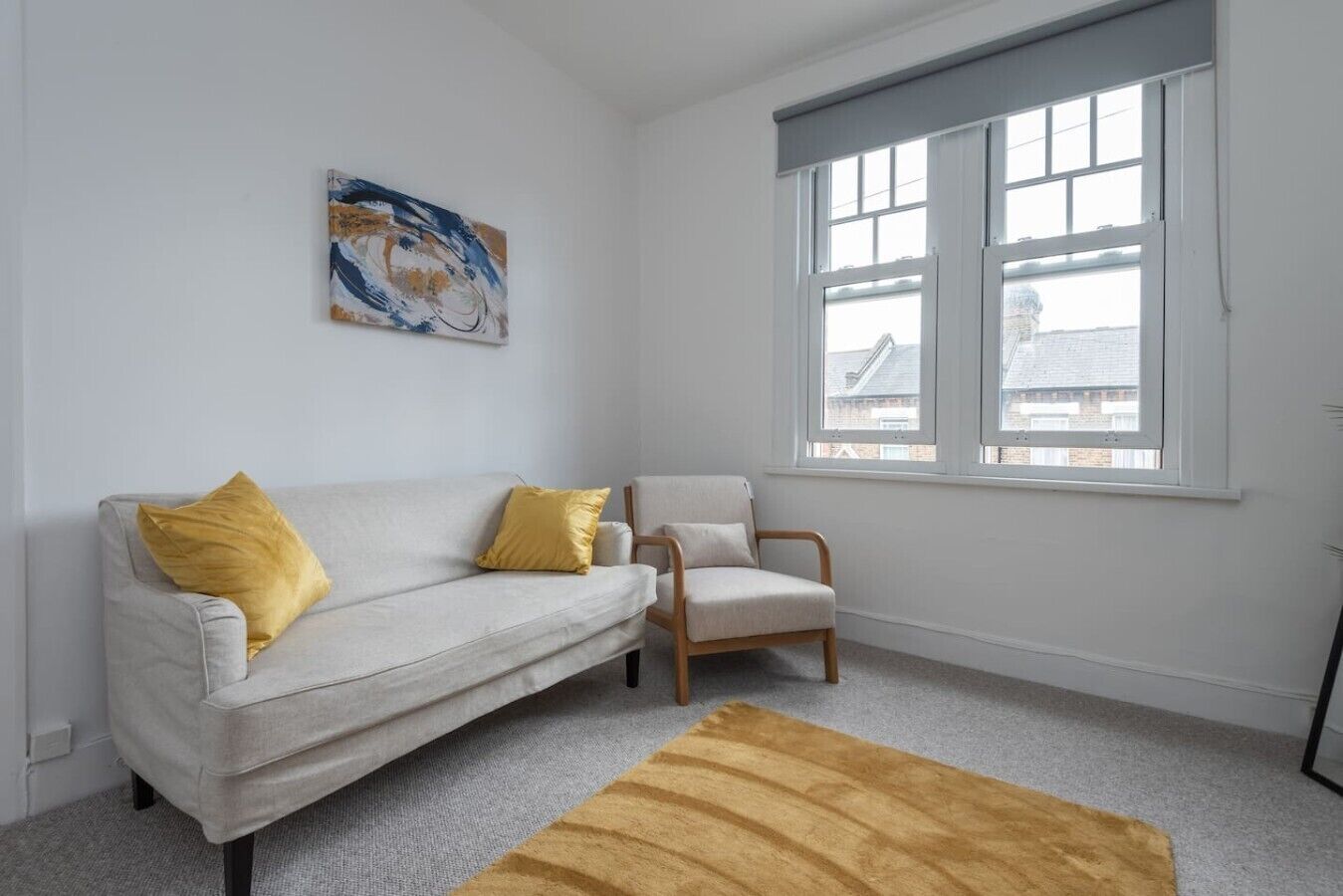 Une pièce avec un canapé blanc, une chaise en bois, un tapis jaune et une fenêtre donnant sur des immeubles.