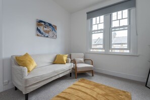 A room with a white sofa, a wooden chair, a yellow rug, and a window with a view of buildings. - Kilburn Flat w/ Designer Kitchen near Kensal Rise (Greater London)