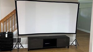 Um quarto com uma grande tela branca de projeção sobre suportes, piso de madeira escura e uma escada ao fundo.