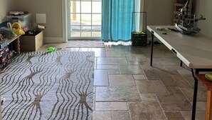Um quarto com uma janela grande, uma porta de correr, um tapete estampado, uma mesa e uma estante com objetos.