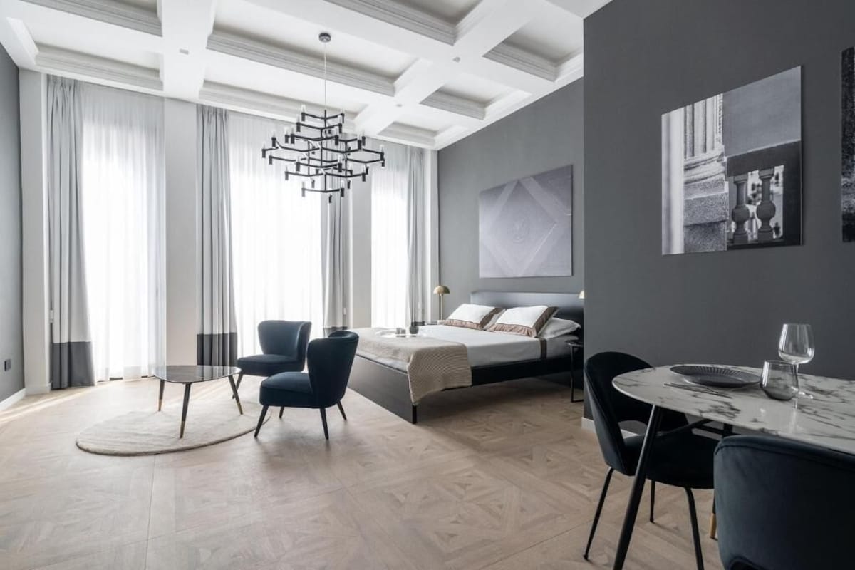 A modern bedroom with a bed, a dining table, and chairs, a large window with curtains, and a hanging light fixture.