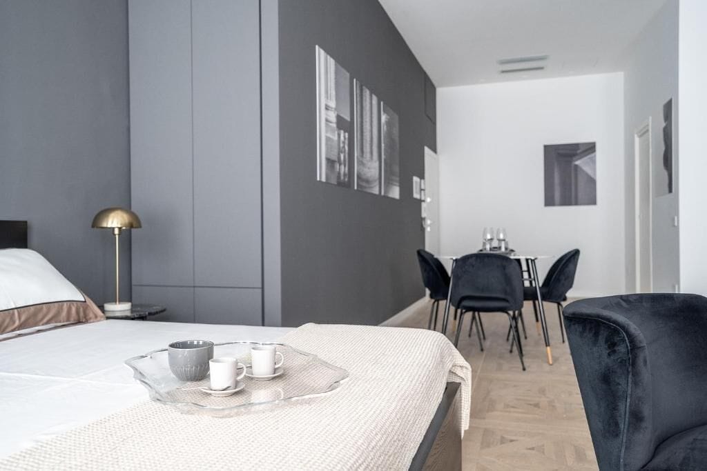 A modern hotel room with a bed, bedside table, lamp, and a dining area with chairs and a table.