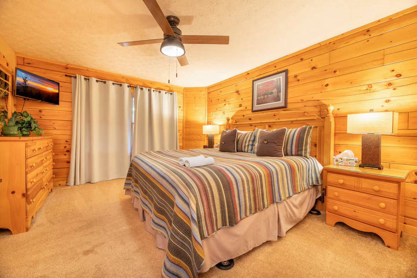 A cozy wooden room with a bed, a ceiling fan, a TV, and a framed picture on the wall.