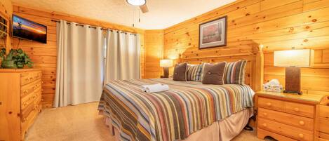 A cozy wooden room with a bed, a ceiling fan, a TV, and a framed picture on the wall.