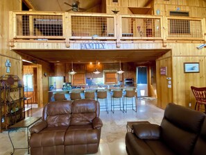 A spacious living area with a high ceiling, a central bar with stools, and a large leather sofa. - Heated Pool, Cabana, Luxury, Mt. Views, 2 minutes 2 Frio River & Garner Park! (Concan)