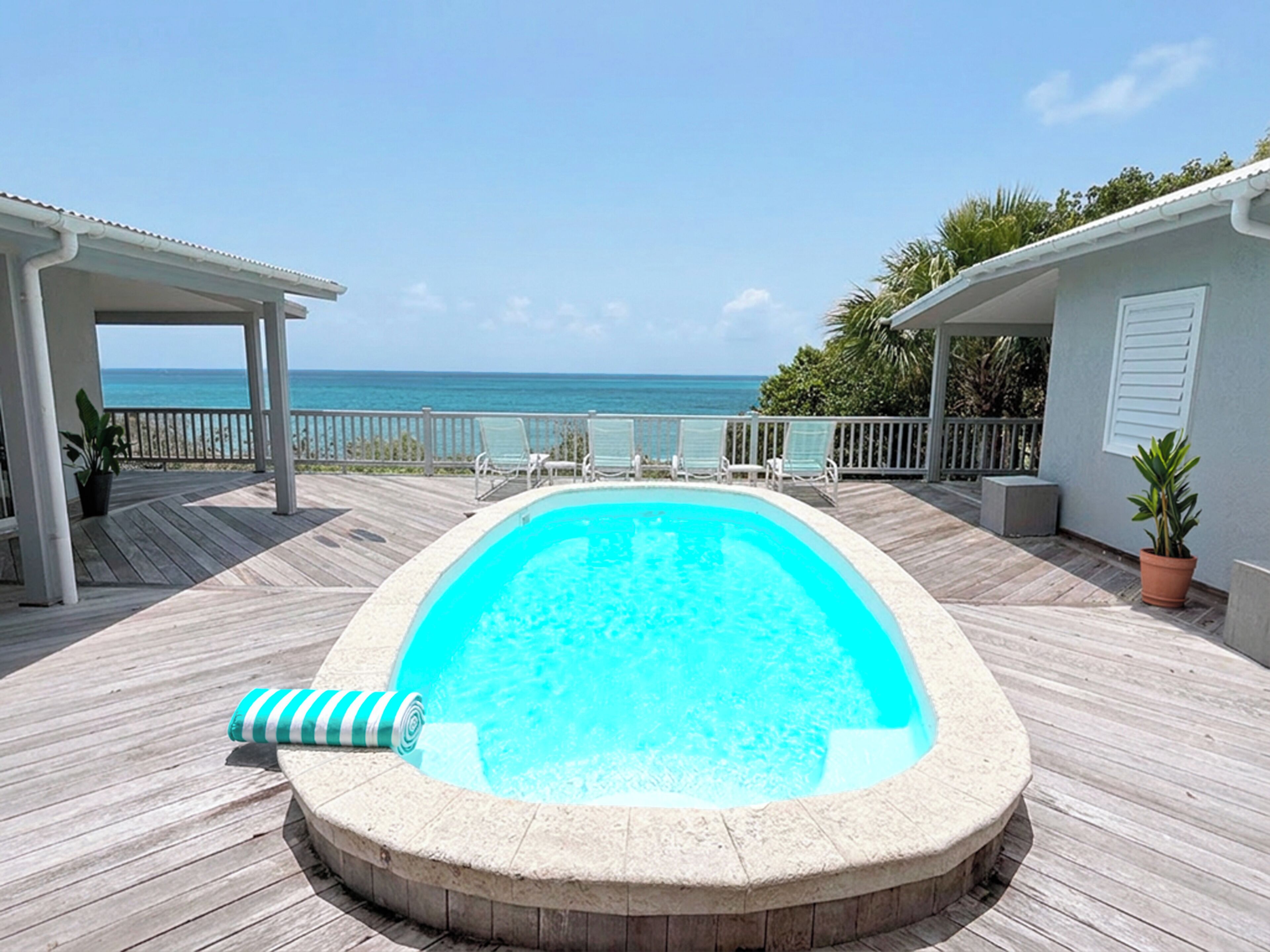A swimming pool with a wooden deck, a lounge area with chairs, and a view of the ocean.