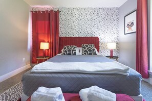 Een hotelkamer met een bed, twee bedlampen, een rood gordijn, een behang met patroon en een ingelijst schilderij aan de muur.