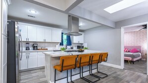 A modern kitchen with white cabinets, a stainless steel refrigerator, a bar area with four stools, and a view into a bedroom with a pink bed. - Outdoor Cinema | Hot Tub | Fire Pit | Huge Yard (Houston)