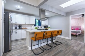 Eine moderne Küche mit weißen Schränken, einem Edelstahlkühlschrank, einer Bar mit vier Hockern und Blick in ein Schlafzimmer mit einem rosafarbenen Bett.