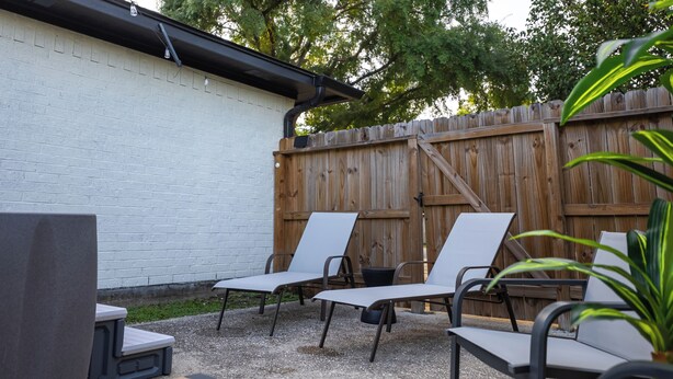 An outdoor patio area with a white brick wall, a wooden fence, and three white lounge chairs. - Outdoor Cinema | Hot Tub | Fire Pit | Huge Yard (Houston)